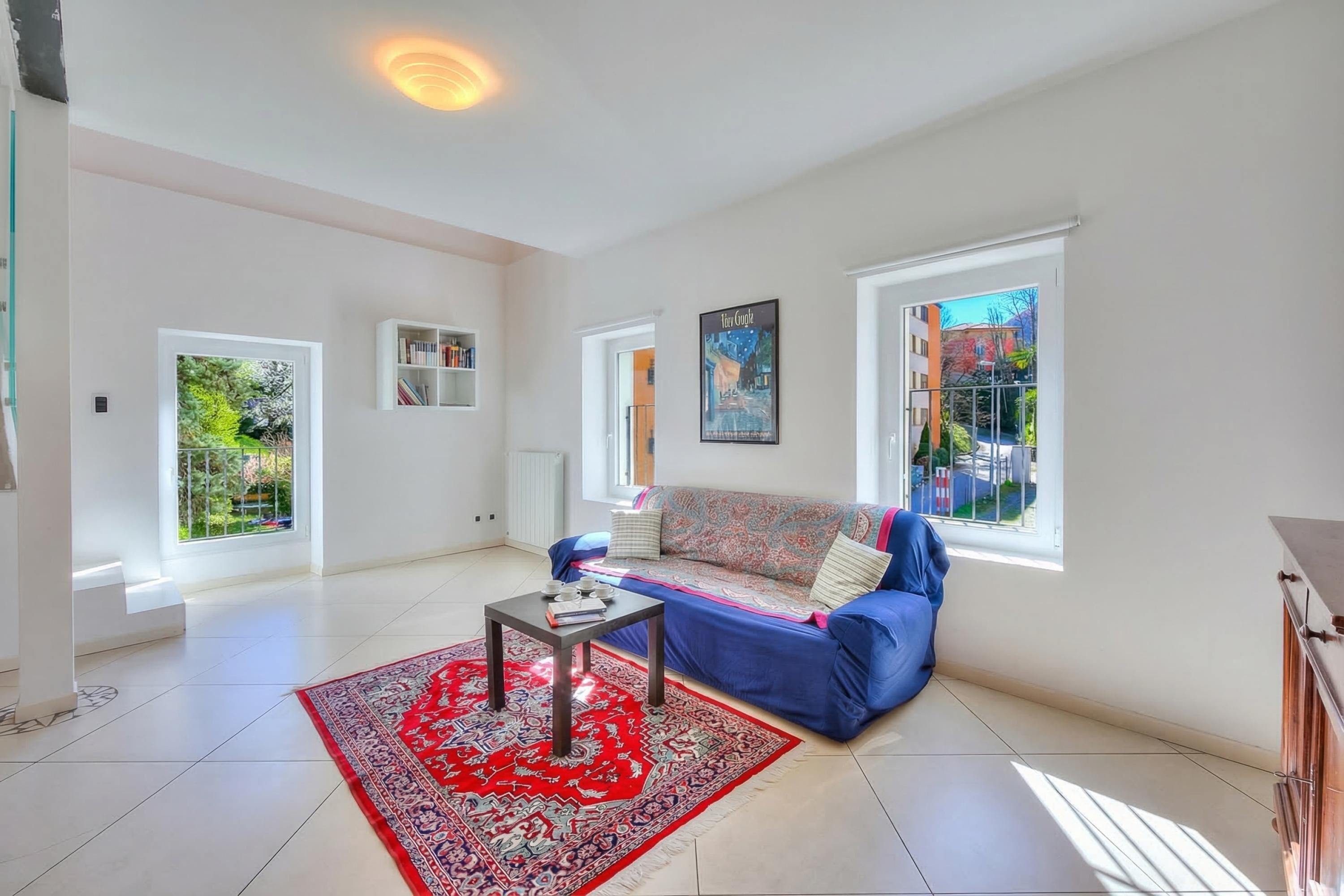 A spacious living room with a blue sofa, a red patterned rug, and a bookshelf.
