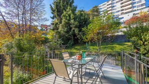 Un balcón con una mesa y sillas de vidrio, rodeado por una baranda metálica, con vistas a un jardín con árboles y un edificio al fondo.