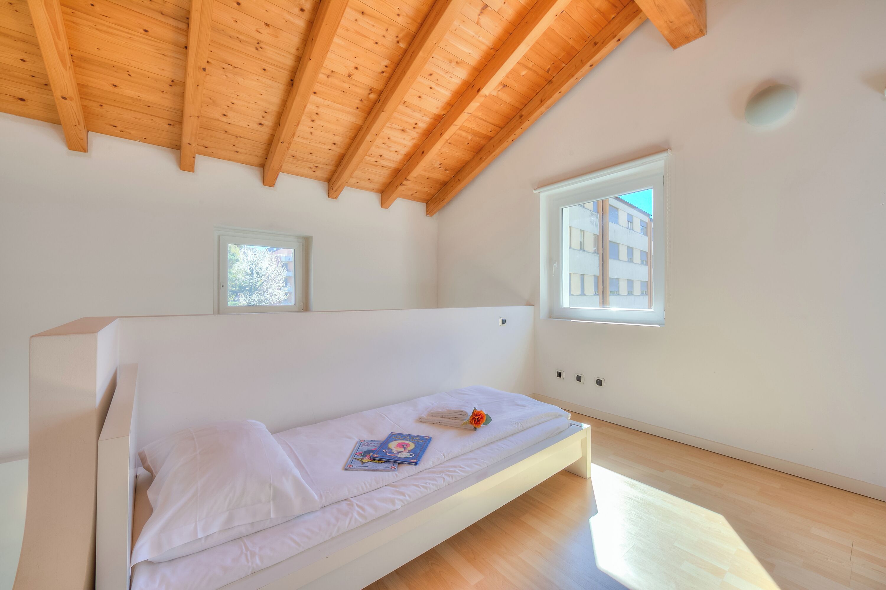 A single bed with a white mattress, a book, and a flower on it. There is a window on the right side of the bed, and a wooden ceiling above.