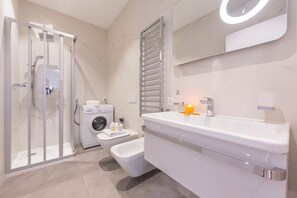 A modern bathroom with a glass-enclosed shower, a built-in washing machine, a bidet, and a large mirror with a circular light.