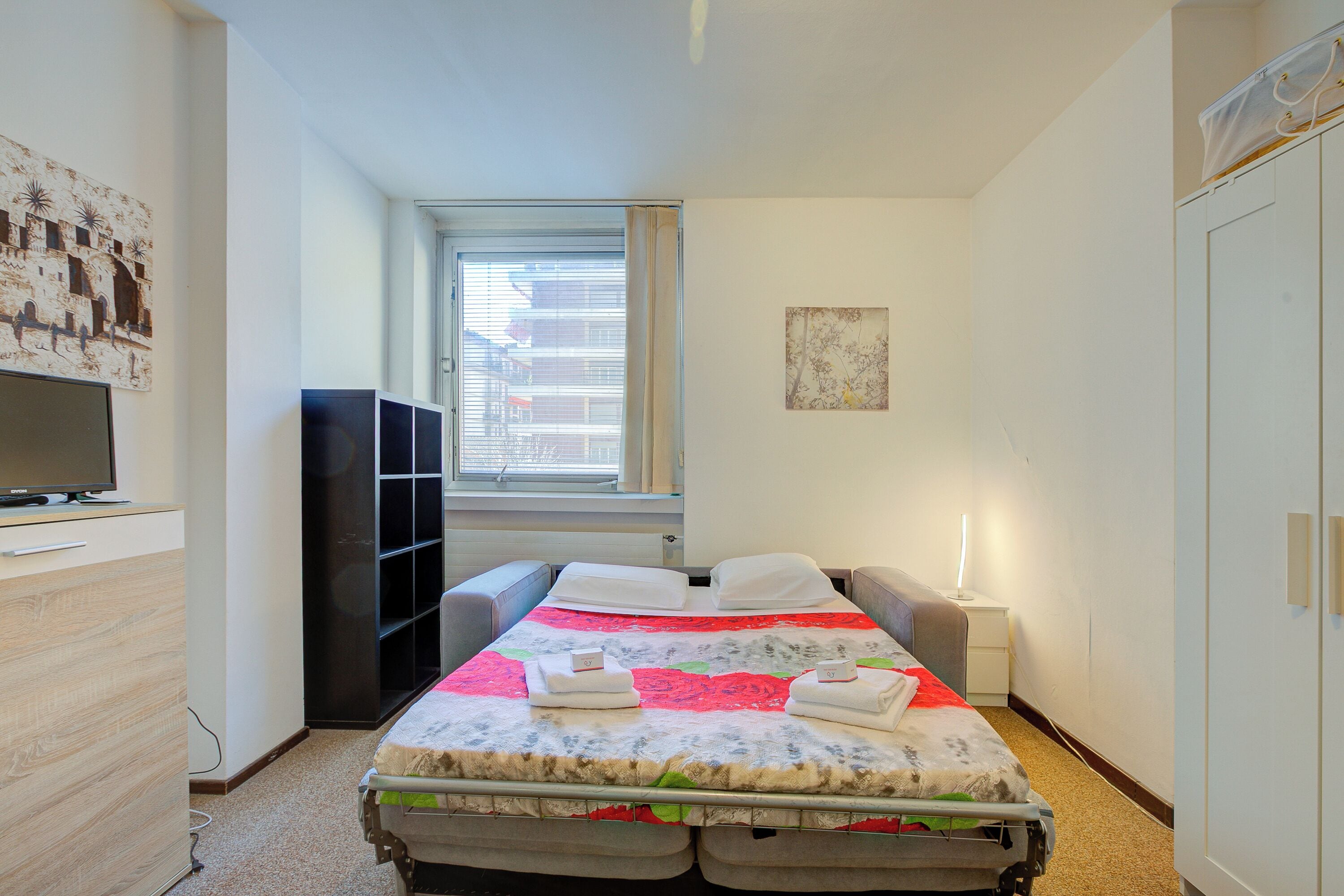 A hotel room with a bed, a TV, a wardrobe, and a window with blinds.