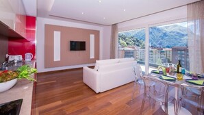 Un salon moderne avec un canapé blanc, un coin repas avec des chaises transparentes, un téléviseur à écran plat fixé au mur et une grande fenêtre offrant une vue sur les montagnes.