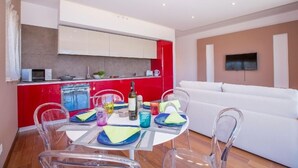 Un salon moderne avec un canapé blanc, une table à manger avec des chaises transparentes et un coin cuisine doté de meubles rouges et blancs.