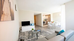 En moderne stue med sofa, sofabord og fladskærms-tv. Der er et spiseområde med bord og stole, og et køkken i baggrunden.