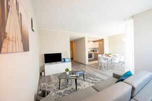 A modern living room with a sofa, a coffee table, and a flat-screen TV. There is a dining area with a table and chairs, and a kitchen in the background.