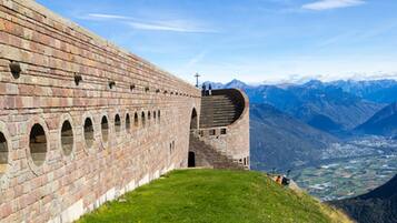 A historic stone wall with arched openings and a cross at the top, overlooking a valley with mountains in the background.