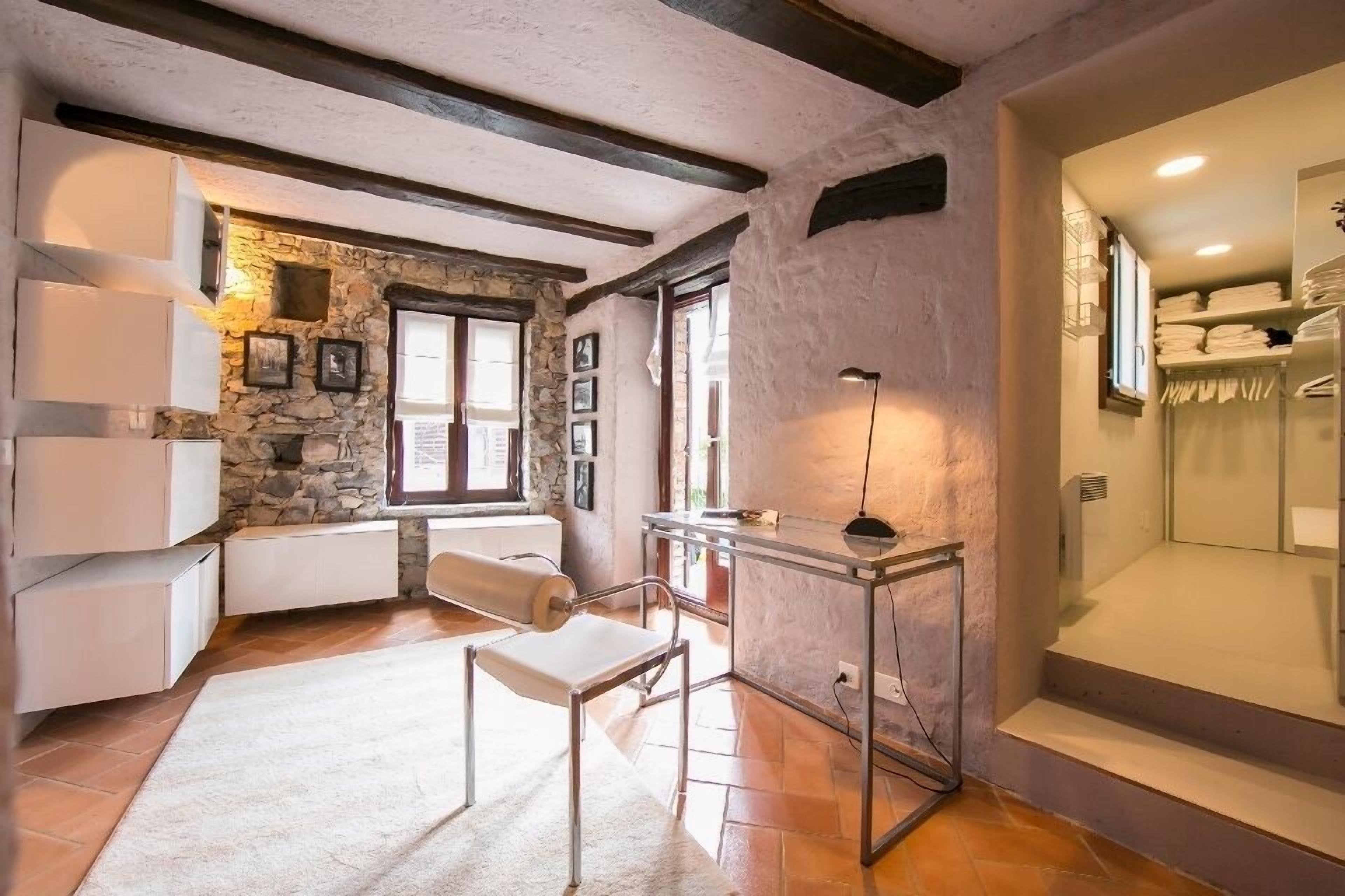 Quarto com parede de pedra, teto de madeira, radiador branco, escrivaninha, cadeira e área de chuveiro envidraçada.