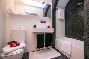 Une salle de bain moderne équipée de toilettes, d’un lavabo et d’une baignoire. On y trouve des serviettes noires suspendues à des crochets, un tapis blanc sur le sol et un espace douche avec un mur en mosaïque.