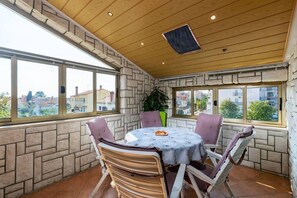 A sunlit patio with a round table and chairs, stone walls, and large windows offering a view of the surrounding neighborhood.