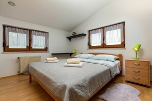 A bedroom with a bed, two windows with curtains, a radiator, a bedside lamp, and a small shelf.