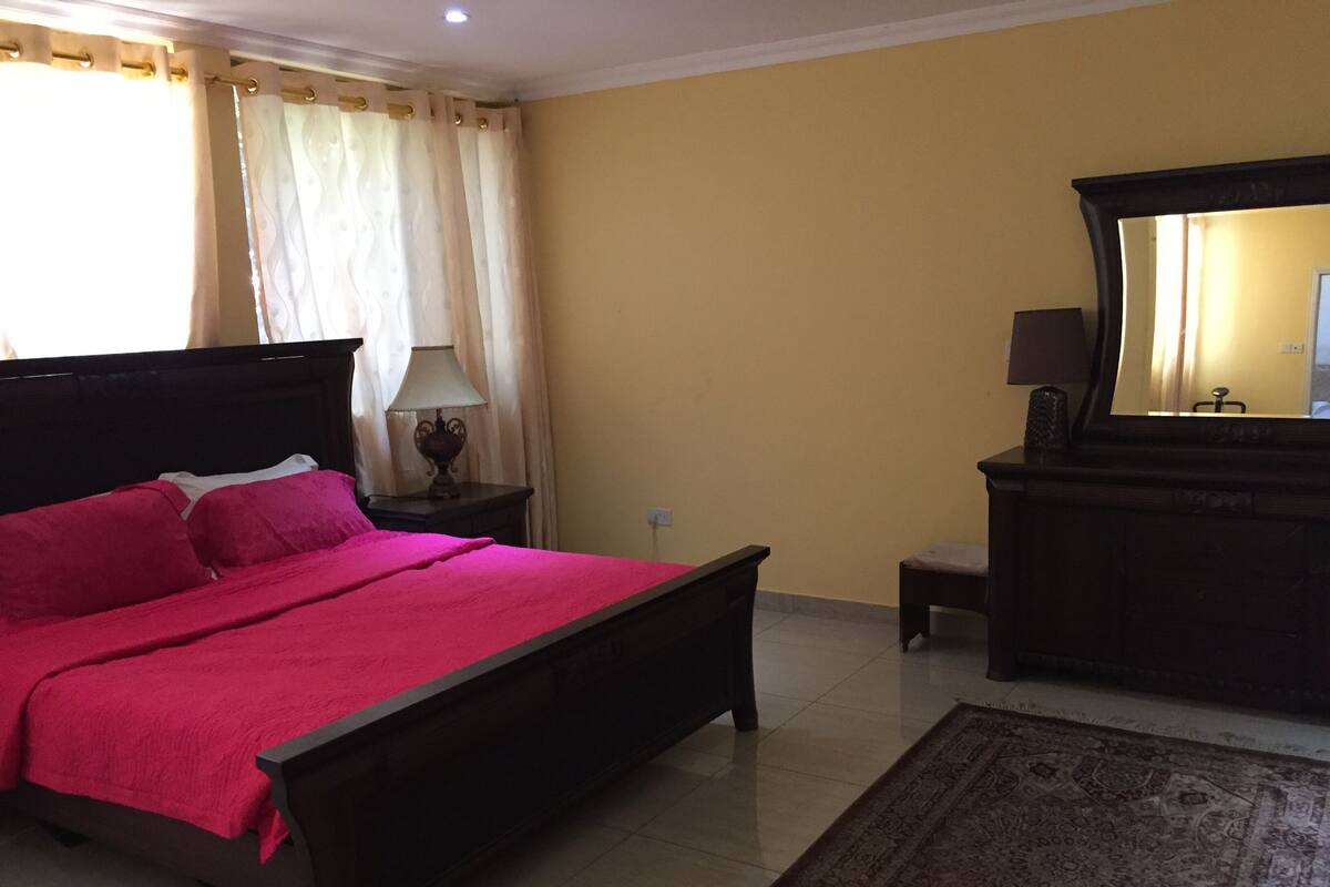 A bedroom with a bed, nightstand, lamp, dresser, mirror, and a rug.