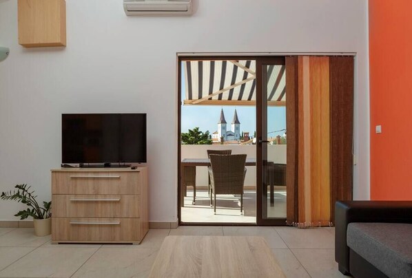 A modern living room with a flat-screen TV on a wooden cabinet, a sliding glass door leading to an outdoor patio with a view of a church and a table with chairs.