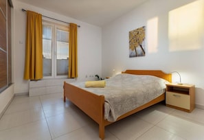 Un dormitorio con cama, mesita de noche, ventana con cortinas y un cuadro colgado en la pared.