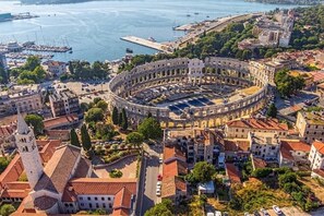 An aerial view of a historic city with a prominent round amphitheater, surrounded by traditional buildings and a waterfront.