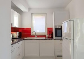 A modern kitchen with white cabinets, a stainless steel refrigerator, a coffee maker, and a window with blinds.
