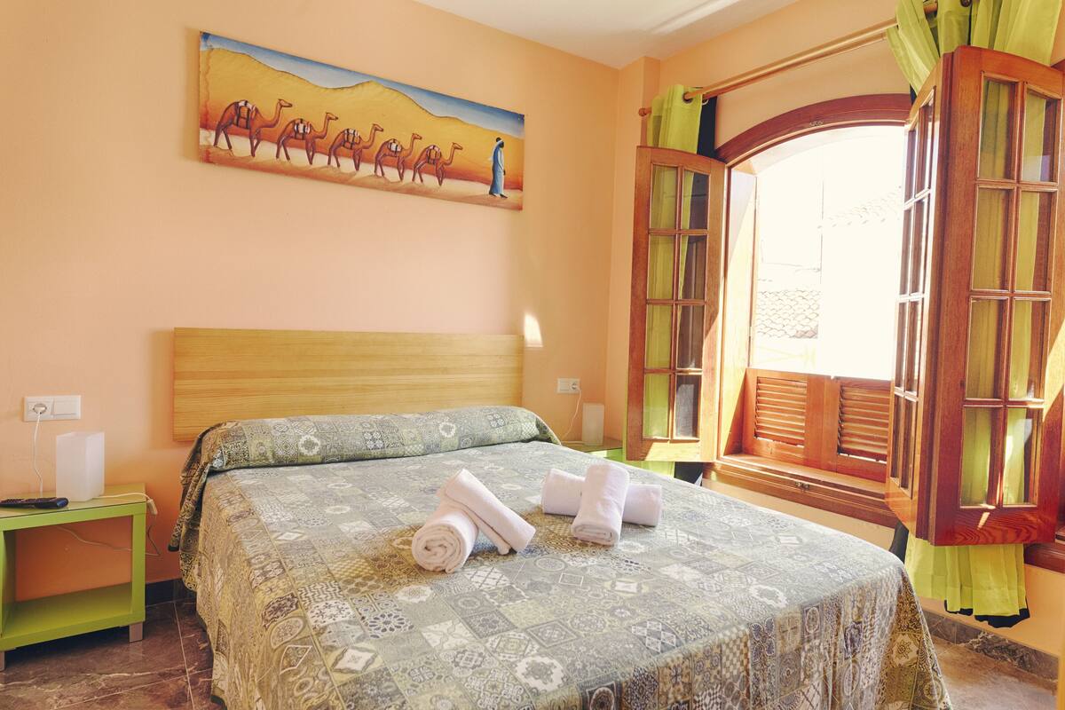 Une chambre d’hôtel avec un lit, une table de chevet, un tableau représentant des chameaux et une fenêtre avec des rideaux.