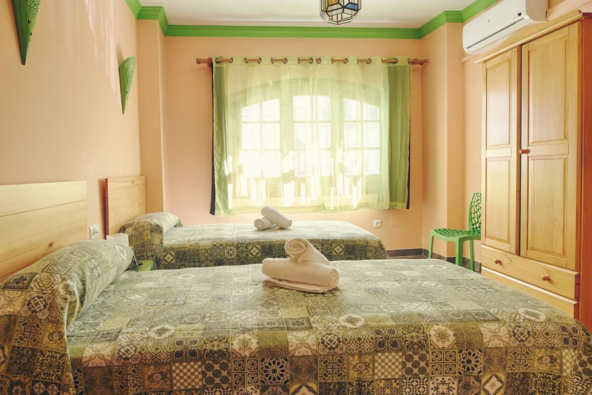 Une chambre avec deux lits, une fenêtre avec des rideaux légers, un fauteuil vert et une armoire.