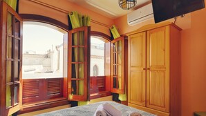 Habitación con cama, dos ventanas con contraventanas abiertas, un armario de madera y vistas a edificios.