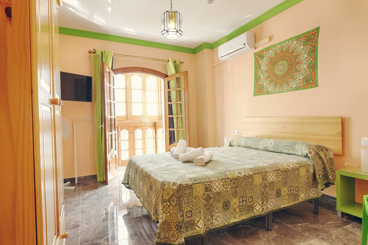 Une chambre à coucher avec un lit, un rideau vert, une tête de lit en bois, une table de chevet verte et un climatiseur fixé au mur.