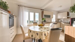 Une cuisine moderne avec des meubles blancs, une table à manger recouverte d’une nappe à motifs floraux et une vue sur l’extérieur grâce à de grandes fenêtres.