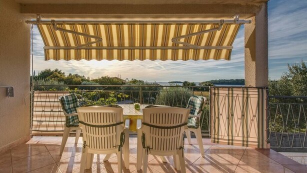 Un balcon avec une table et des chaises, un auvent à rayures et une vue sur la verdure et l’eau.