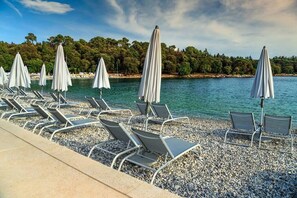 A pebbled beach with rows of sun loungers and umbrellas, overlooking a calm sea and a forested coastline. - Villa Tanga near Rovinj for 8 persons with pool (Rovinj)