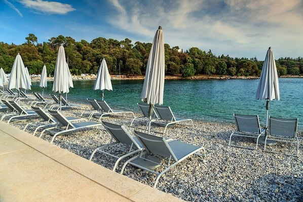 A beachside area with rows of sun loungers and umbrellas, a pebble beach, and a calm sea with a forested shoreline in the background. - Villa Tanga near Rovinj for 8 persons with pool (Rovinj)