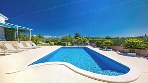 Une piscine rectangulaire à bord incurvé, entourée d’une terrasse carrelée et d’un espace de détente couvert équipé de chaises longues.