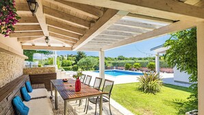 Une terrasse couverte avec une table et des chaises en bois, un sol en pierre et une vue sur une piscine et la verdure.