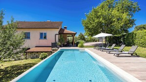 A rectangular swimming pool with a tiled deck, a stone wall, and a house with a tiled roof.