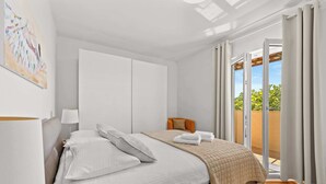 A bedroom with a bed, a nightstand, a lamp, a window with curtains, and a view of greenery outside.