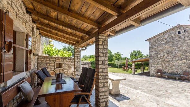 Un espacio de comedor al aire libre cubierto, con paredes de piedra, vigas de madera y vistas a un jardín y un edificio al fondo.