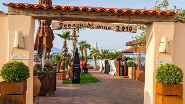 A wooden archway with the text "Seensicht anno 2010" and a pathway leading to a beach area with palm trees and outdoor seating.
