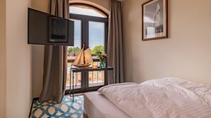 A hotel room with a bed, a TV mounted on the wall, a window with curtains, a small table with a lamp and flowers, and a framed picture on the wall.