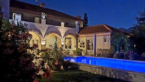 Un área junto a la piscina con piscina, plantas en macetas y un edificio con ventanales arqueados.