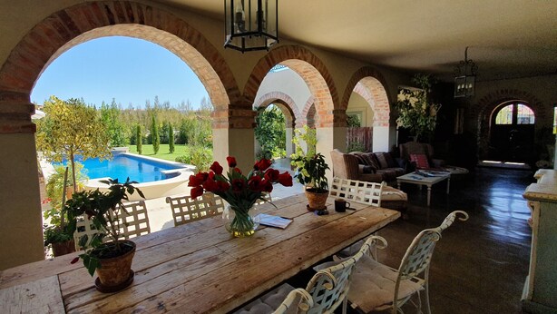 Un patio cubierto con piscina, mesa de madera, plantas en macetas y mobiliario exterior.