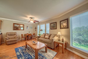 A living room with a sofa, coffee table, and a ceiling fan. There are windows with blinds, a dining area with chairs, and a framed picture on the wall.