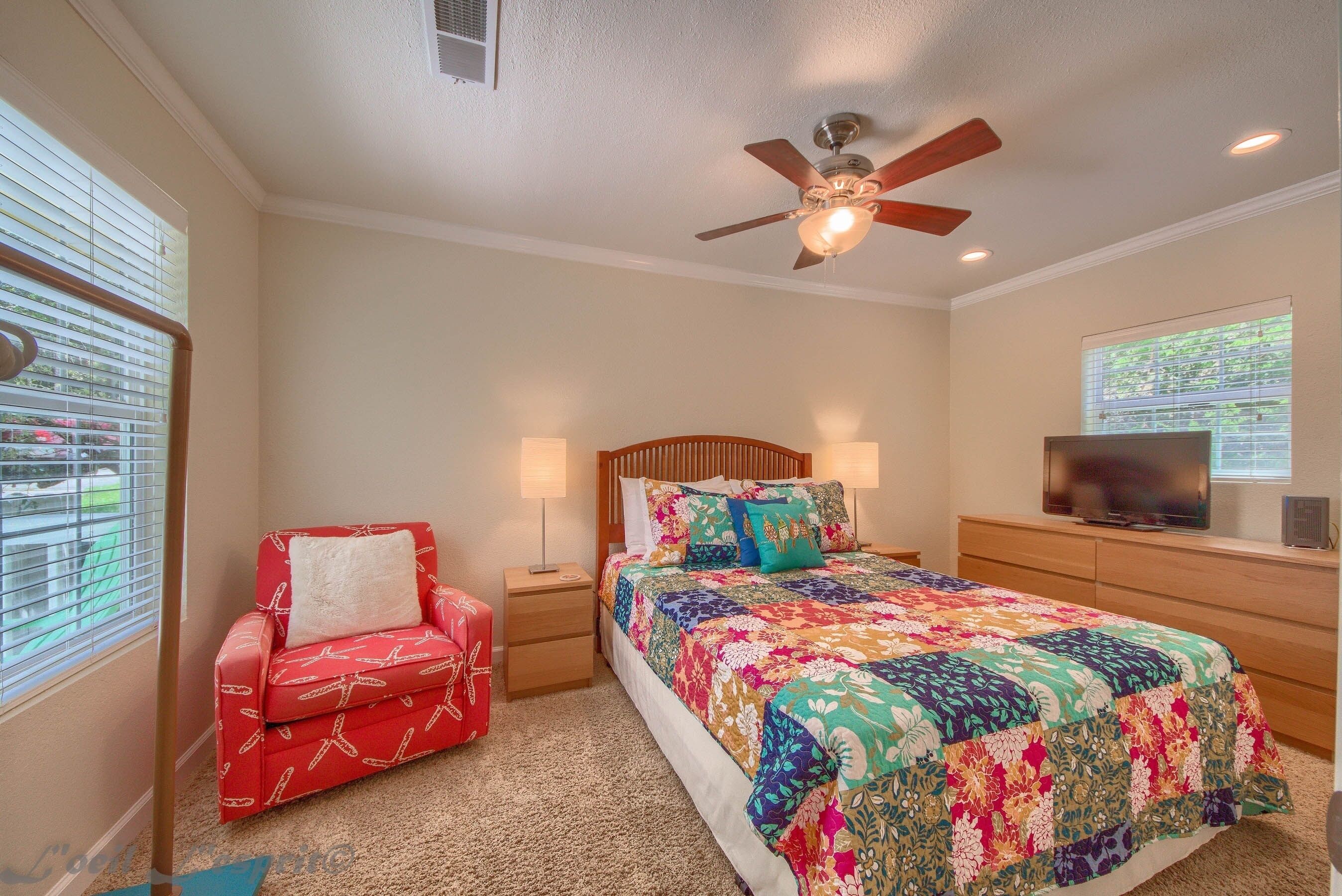 A bedroom with a bed, a red chair, a ceiling fan, a TV, and a window with blinds.