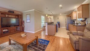 A spacious living area with a wooden entertainment center, a large flat-screen TV, and a comfortable leather sofa. A kitchen with wooden cabinets and a refrigerator is visible in the background.