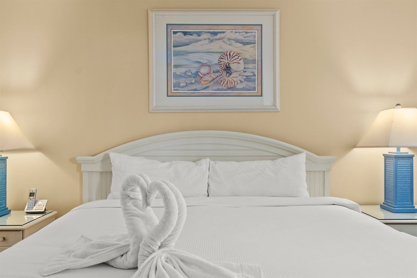 A neatly made bed with white linens, a framed artwork above it, and two bedside lamps.