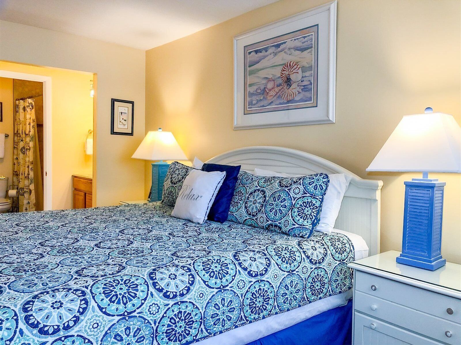 A bedroom with a bed, two bedside lamps, a framed picture on the wall, and a bathroom visible through an open door.
