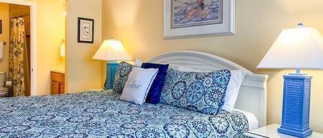 A bedroom with a bed, two bedside lamps, a framed picture on the wall, and a bathroom visible through an open door.