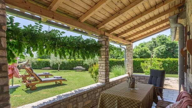 Ein überdachter Patio-Bereich mit Tisch und Stühlen, eine Steinsäule und Blick auf einen gepflegten Garten.