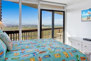 A bedroom with a large bed, a white dresser, and a view of the beach through a sliding glass door.