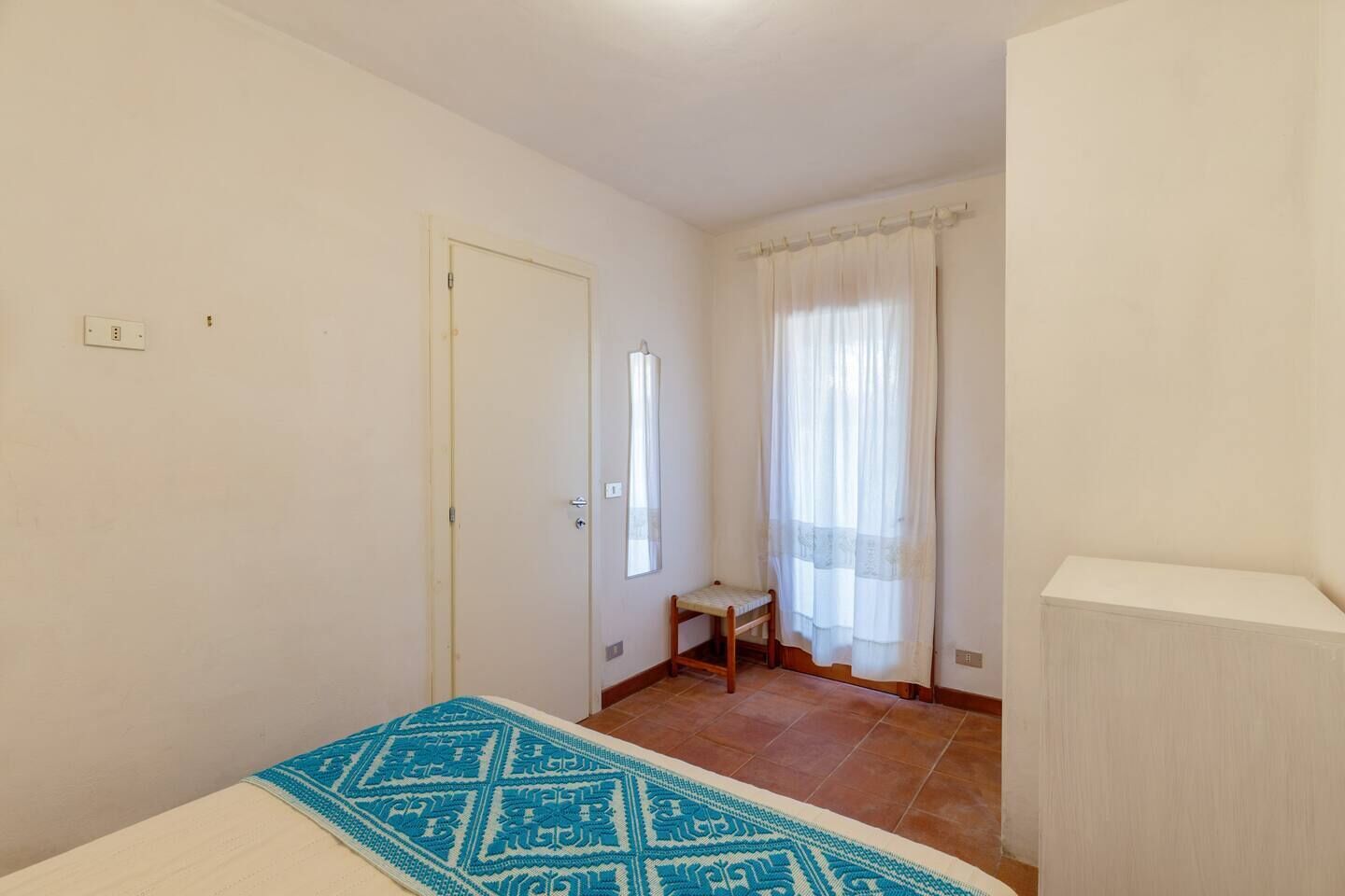 Ein kleiner, sauberer Raum mit einem Bett, einem Sessel, einem Fenster mit Vorhängen und einer Tür.
