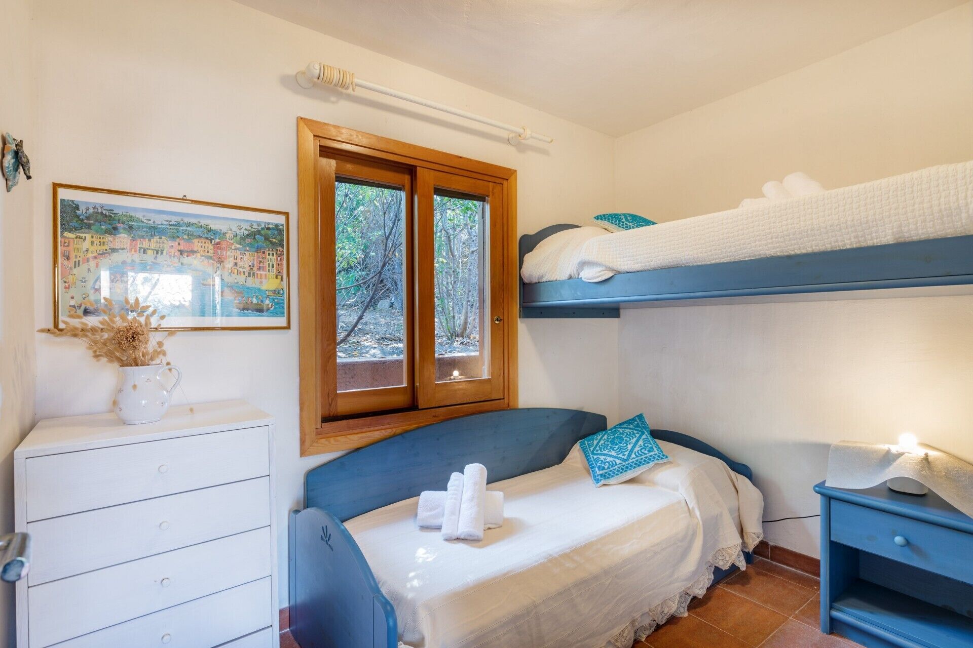 A bedroom with a bunk bed, a bedside table with a lamp, a dresser, and a window with a view of trees.