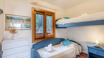 A bedroom with a bunk bed, a bedside table with a lamp, a dresser, and a window with a view of trees.