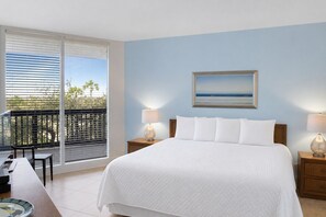 A hotel room with a bed, two bedside tables with lamps, a painting of a beach, a balcony with a table and chairs, and a view of palm trees.