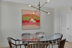 A dining area with a glass table surrounded by wicker chairs, a modern chandelier, and a large abstract painting on the wall.