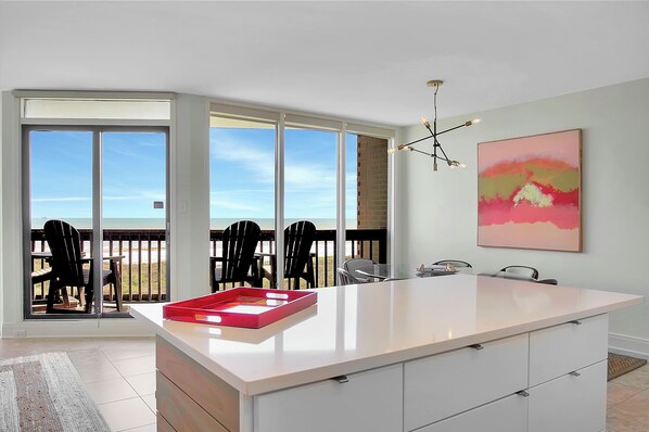 A modern kitchen with a large island, a dining area with chairs, and a view of the beach through large sliding doors.